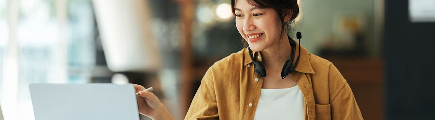 A woman wearing a brown shirt smiles while working on a laptop. She has headphones around her neck and is sitting in a cozy indoor setting.