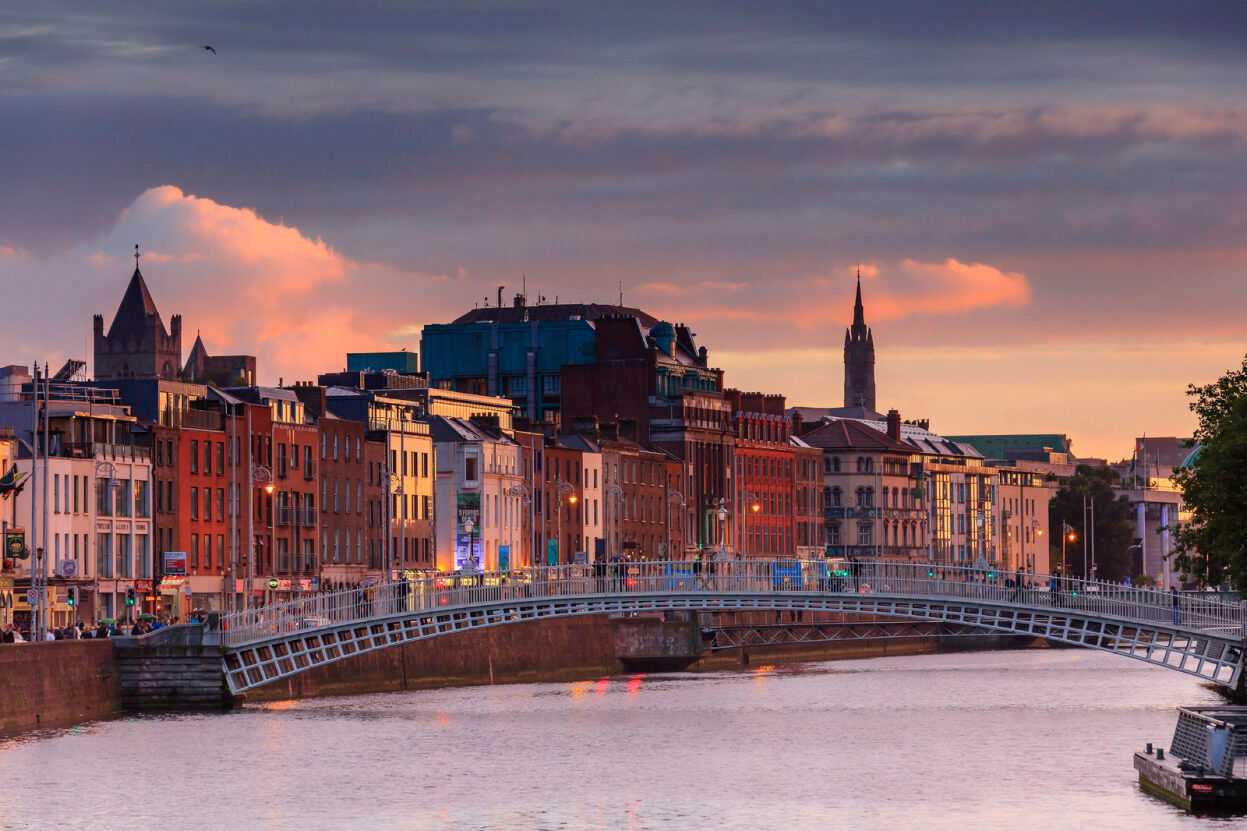A view of colorful riverside buildings and the Hapenny Bridge over the River Liffey in Dublin, Ireland at sunset, with a dramatic sky and church spires in the background.