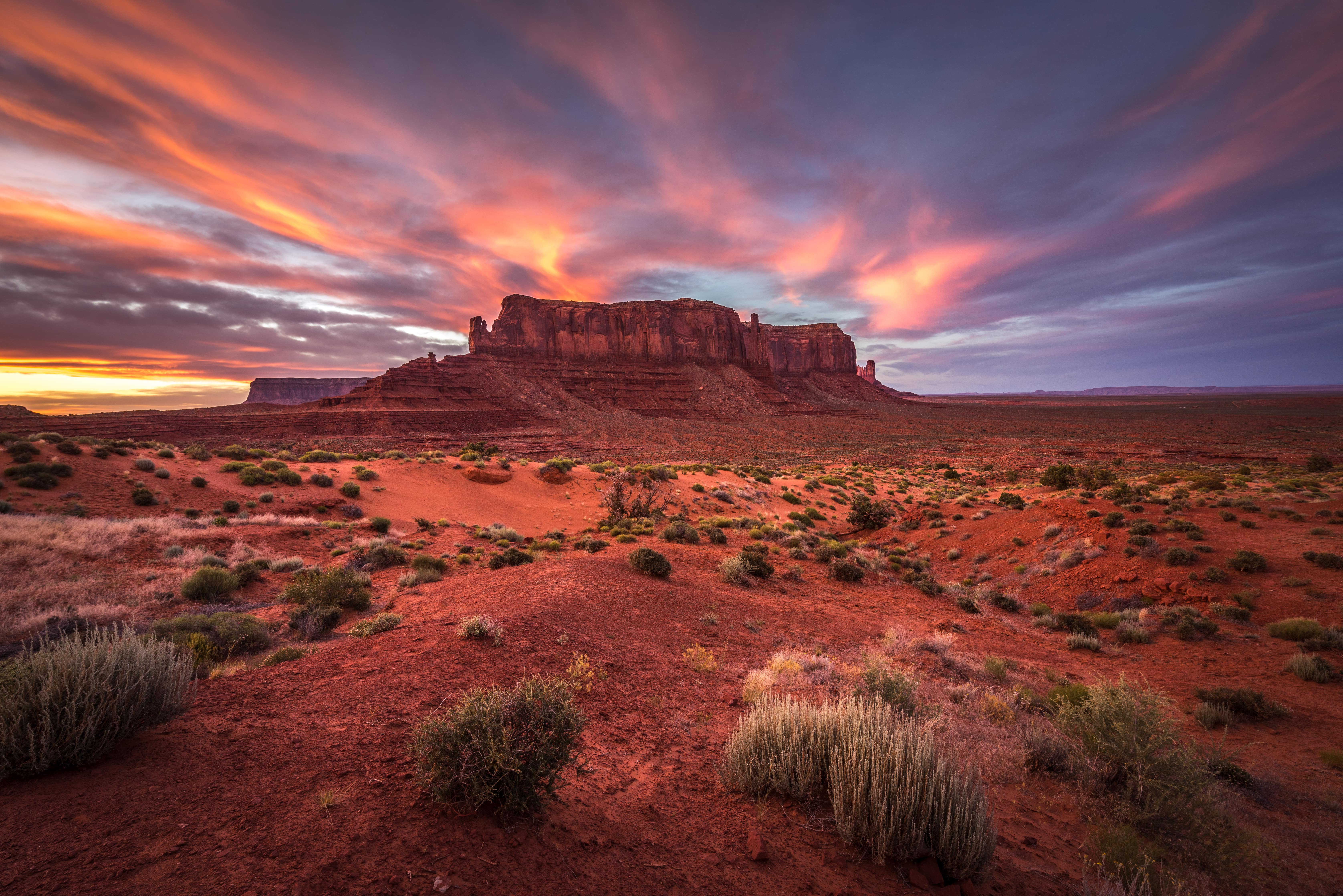 A breathtaking desert landscape at sunset, featuring a large sandstone butte surrounded by red sand and sparse vegetation. The sky is ablaze with vibrant orange, pink, and purple clouds, casting a warm glow over the scene.